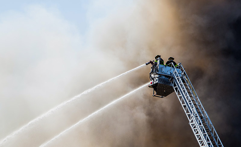 Deux pompiers sur une nacelle arrosent de l’eau dans une épaisse fumée en combattant un grand incendie.