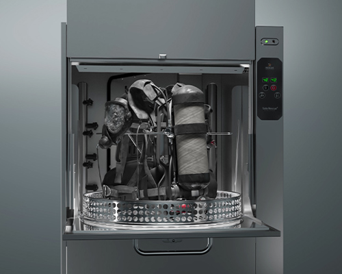 Open industrial washing machine with firefighter gear inside, including a breathing apparatus; control panel on the right side.