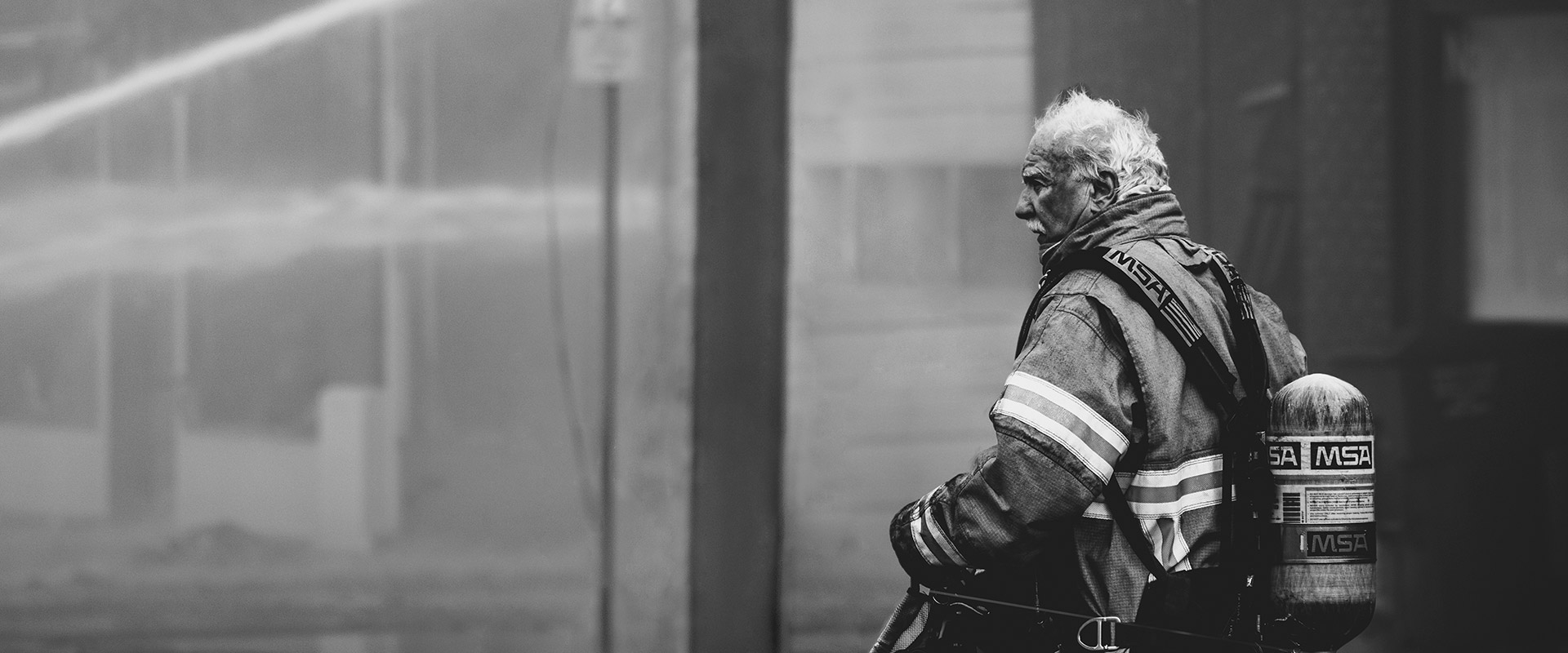 Schwarzweißfoto eines Feuerwehrmanns in kompletter Ausrüstung mit „MSA“-Sauerstoffflasche, stehend vor einem verrauchten Gebäude an einem Brandort.