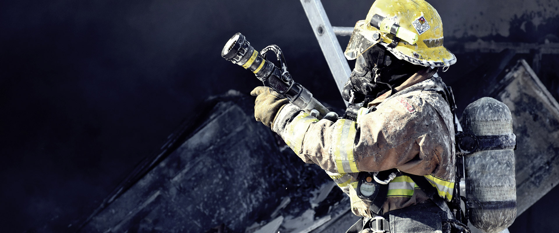 Feuerwehrmann in kompletter Ausrüstung mit Helm und Atemschutzgerät, richtet den Schlauch in einer rauchigen, dunklen Brandszene, Uniform mit Ruß bedeckt.