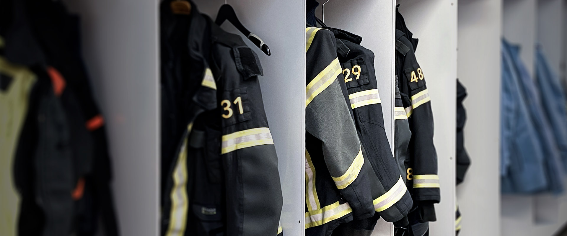 Vestes noires de pompiers avec des bandes jaunes et les numéros 31, 29 et 48 suspendues dans une rangée de casiers.