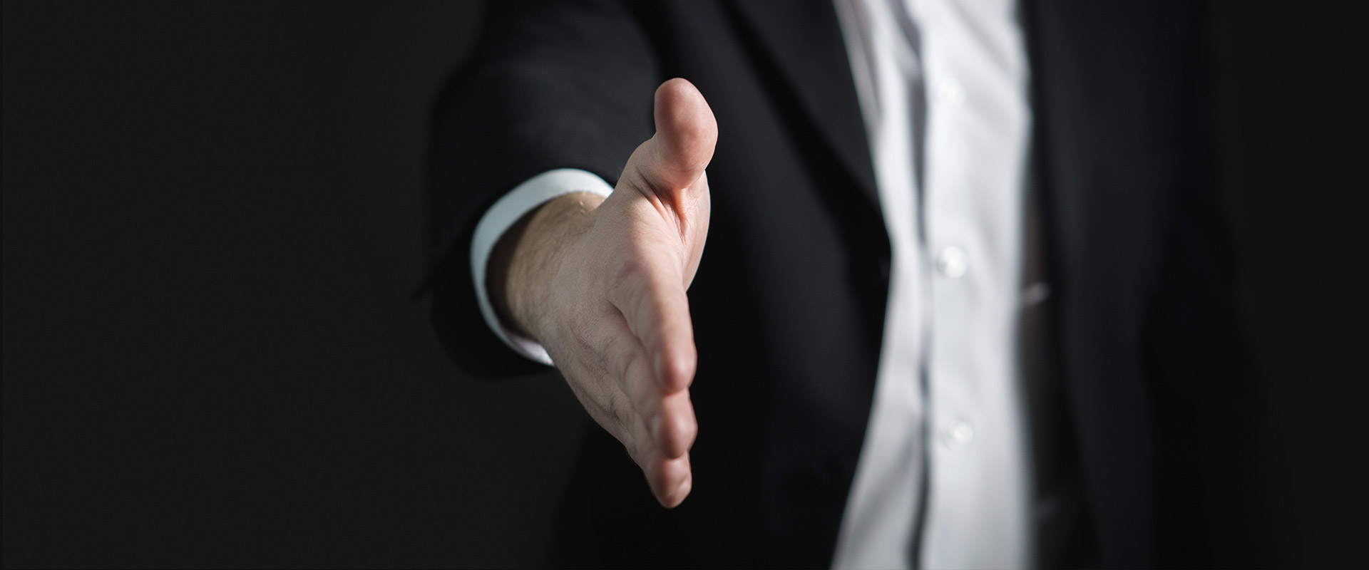 Person i svart kostym och vit skjorta som sträcker ut höger hand för ett handslag mot en mörk bakgrund.